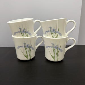 May include: Four white ceramic mugs stacked in pairs. Each mug features a blue iris flower design with green stems and leaves. The mugs have a curved handle and a smooth, glossy finish, suitable for beverages.