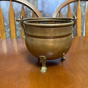 May include: A small copper pot with a handle and three feet. The pot is sitting on a wooden table.