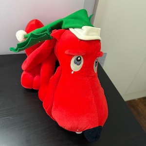May include: A plush red creature with large, expressive eyes and a black beak. It wears a green elf hat with a white brim and a small white pom-pom. The toy is made of soft, fuzzy material.
