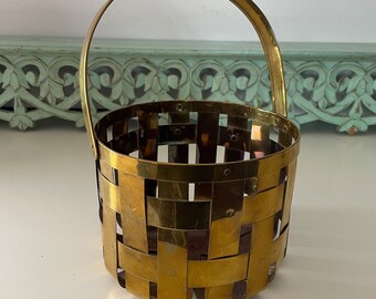 May include: A small, gold-toned, woven metal basket with a handle. The basket is made of thin strips of metal that are woven together to create a lattice pattern.