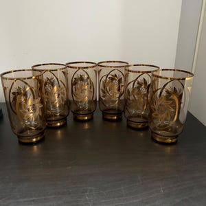 Peut inclure: Ensemble de sept verres à grand gobelet vintage. Chaque verre présente un corps transparent avec un bord doré et un motif floral doré. Les verres sont disposés sur une surface sombre. Un ajout classique à toute collection de bar.