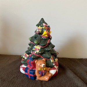 May include: A ceramic Christmas tree figurine with a teddy bear sitting at the base. The tree is decorated with red and yellow ribbon, green garland, and gold bells. The teddy bear is wearing a red and blue bow tie.