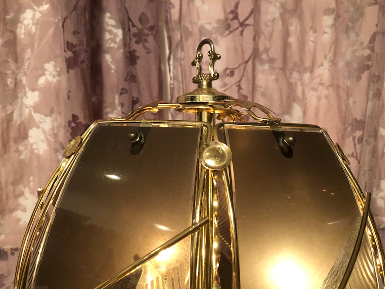 May include: A gold-toned metal lampshade with a geometric design. The lampshade has a frosted glass panel and a decorative finial on top.