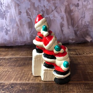 May include: A ceramic Christmas figurine of three Santas stacked on top of each other. The Santas are wearing red and white suits with black boots and green trim. The figurine is sitting on a white base.