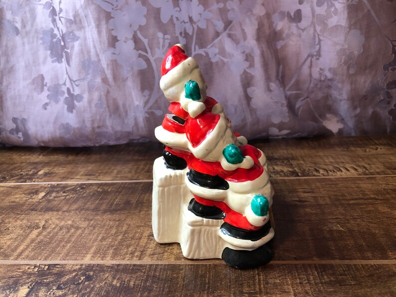 May include: A ceramic Christmas figurine of three Santas stacked on top of each other. The Santas are wearing red and white suits with black boots and green trim. The figurine is sitting on a white base.