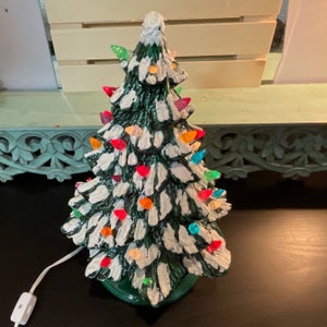 May include: A ceramic Christmas tree with a green base and white snow-like frosting. The tree is decorated with colourful light bulbs, including red, green, yellow, orange, blue, and purple.