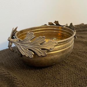 May include: A hammered brass bowl with leaf-shaped handles. The bowl has a shiny, gold finish and is perfect for serving snacks or holding decorative items.