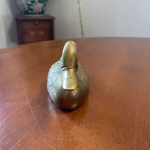 Puede incluir: Una figurita de pato de lat&oacute;n con marcas detalladas de plumas. El pato est&aacute; sentado sobre una superficie de madera.