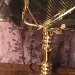 May include: A gold-toned table lamp with a clear glass shade featuring a geometric pattern. The lamp has a round base and a decorative, fluted column.