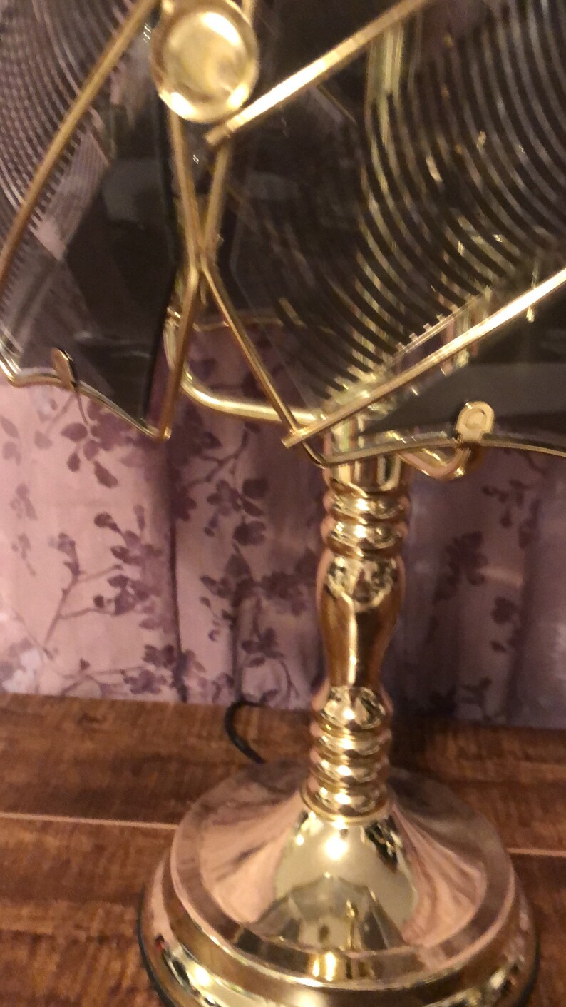 May include: A gold-toned table lamp with a clear glass shade featuring a geometric pattern. The lamp has a round base and a decorative, fluted column.