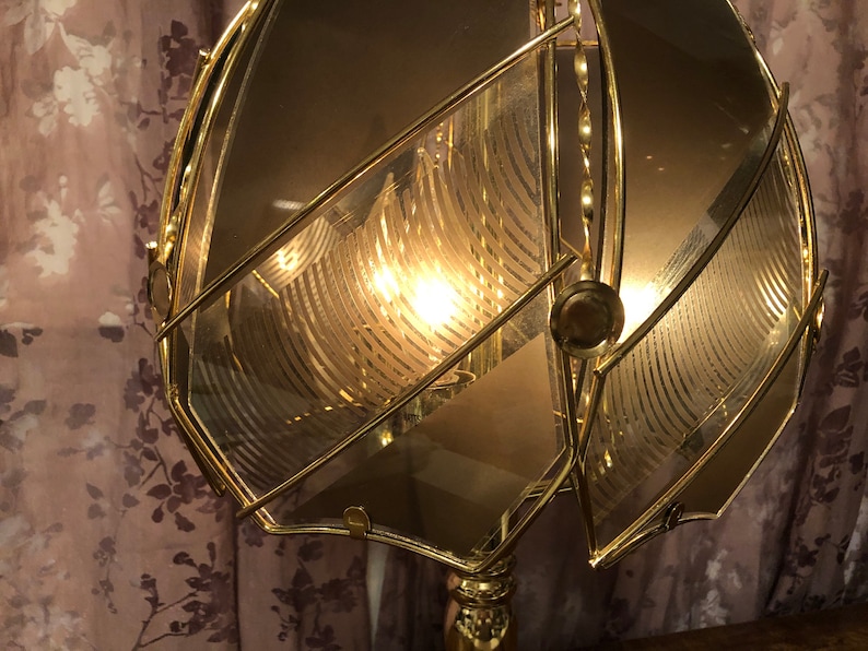 May include: A gold-toned metal lampshade with a geometric design. The lampshade is made of multiple pieces of glass that are joined together to create a unique and intricate pattern. The lampshade is illuminated from within, creating a warm and inviting glow.