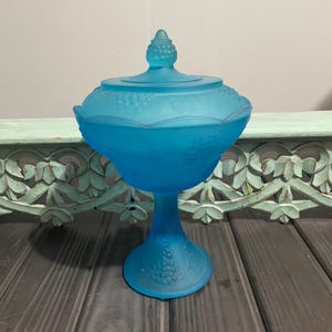May include: A blue glass candy dish with a grape design. The dish has a pedestal base and a lid with a knob on top.