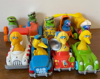 May include: A collection of Sesame Street character toy cars. The cars are yellow, red, blue, green, and white. Each car features a different Sesame Street character, including Big Bird, Cookie Monster, Elmo, and Bert and Ernie.