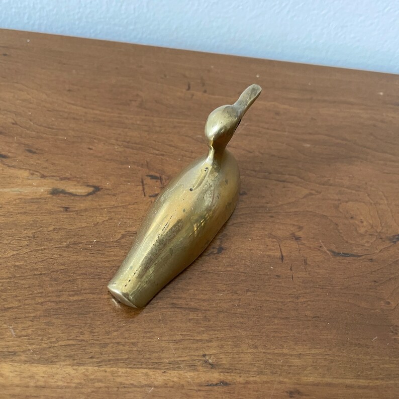 May include: A small, solid brass figurine of a bird. The bird is in a resting position with its head tilted to the side. The figurine is a light gold color.