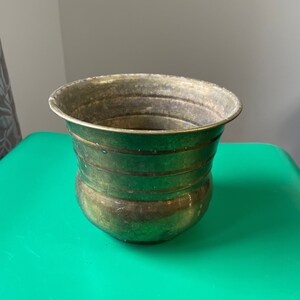May include: A brass planter with a rounded bottom and a slightly flared top. The planter has a worn, aged look and is decorated with horizontal lines.