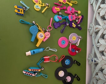 May include: A collection of colorful plastic toys, including musical instruments, whistles, and small records. The toys are attached to keychains and feature various designs, such as musical notes, guitars, and records.