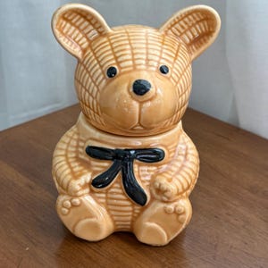 Peut inclure: Un pot à biscuits en céramique beige en forme d'ours en peluche avec un nœud papillon noir. L'ours a une texture de tissage en panier et des yeux et un nez noirs. Le couvercle est posé sur la tête de l'ours.
