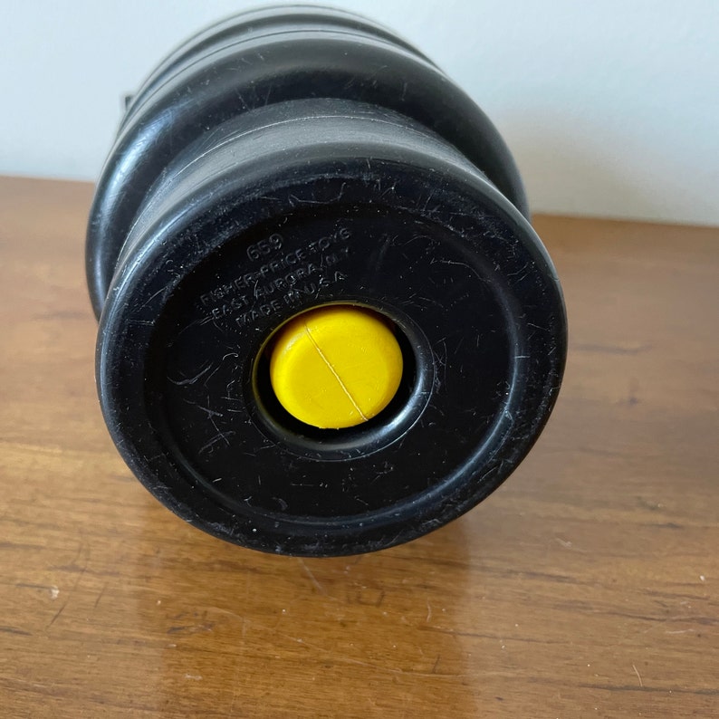 K&ouml;nnte beinhalten: Schwarzes Plastikspielzeug mit einem gelben Knopf. Das Spielzeug hat den Text "659 FISHER-PRICE TOYS EAST AURORA, NY MADE IN U.S.A." auf der Unterseite.