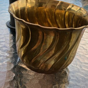 May include: A gold-coloured brass planter with a wavy, textured design. The planter is empty and sitting on a glass table.