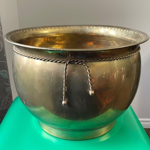 May include: A large, round, brass planter with a rope trim and decorative tassels. The planter is sitting on a green surface.