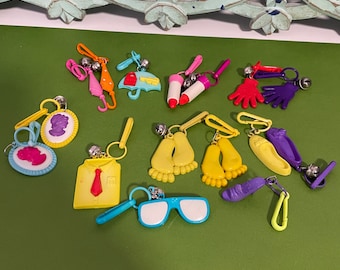 May include: A collection of 12 colorful plastic keychains with various charms, including an umbrella, a lipstick, a pair of feet, a shirt, and a pair of glasses. Each keychain has a small bell attached.