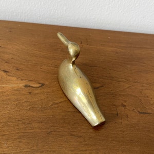 May include: A gold-colored brass bird figurine with a long, slender body and a curved beak. The bird is sitting on a wooden surface.