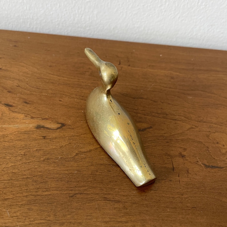 May include: A gold-colored brass bird figurine with a long, slender body and a curved beak. The bird is sitting on a wooden surface.