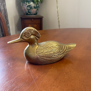 Puede incluir: Una figurita de pato de lat&oacute;n de color dorado con patrones de plumas detallados. El pato tiene un pico largo y una cabeza crestada.