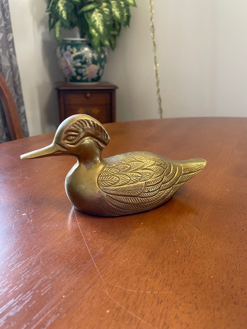 Puede incluir: Una figurita de pato de lat&oacute;n de color dorado con patrones de plumas detallados. El pato tiene un pico largo y una cabeza crestada.