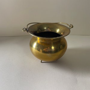 May include: A small, gold-colored brass pot with a handle and three legs. The pot is round and has a smooth, shiny surface.