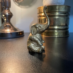 May include: A small, brass elephant figurine with its trunk raised in the air. The elephant is sitting on a dark surface.