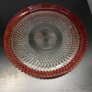 May include: A clear glass serving plate with a red-orange rim and a diamond pattern. The centre of the plate has a starburst design. The plate is round and appears to be vintage.
