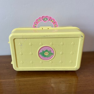 May include: A yellow plastic toy carrying case with a pink handle and a green and yellow sticker of a smiling face with a heart on the front.
