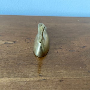 May include: A small, solid brass duck figurine. The duck is in a sitting position with its head tilted back. The duck is a simple, minimalist design.