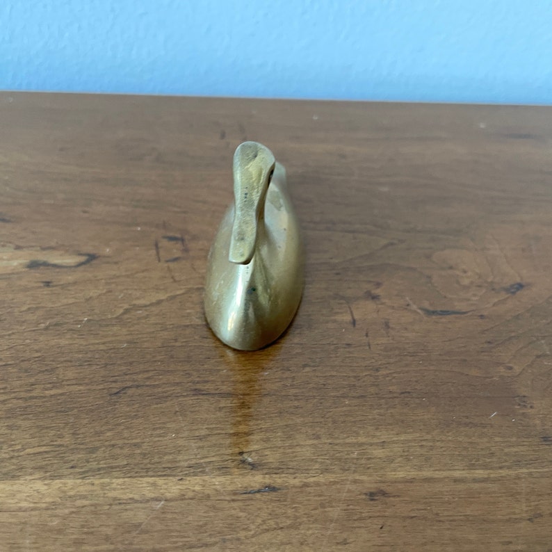 May include: A small, solid brass duck figurine. The duck is in a sitting position with its head tilted back. The duck is a simple, minimalist design.
