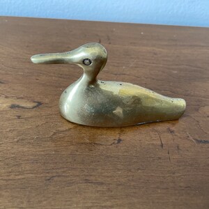 May include: A small, solid brass duck figurine. The duck is sitting with its head turned to the side and its beak slightly open. The duck is a light gold color.