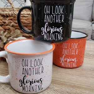 May include: Three ceramic mugs with the text "OH LOOK ANOTHER glorious MORNING". The mugs are black, orange, and white with speckled designs. The white mug has an orange rim, and the black mug has a black handle.
