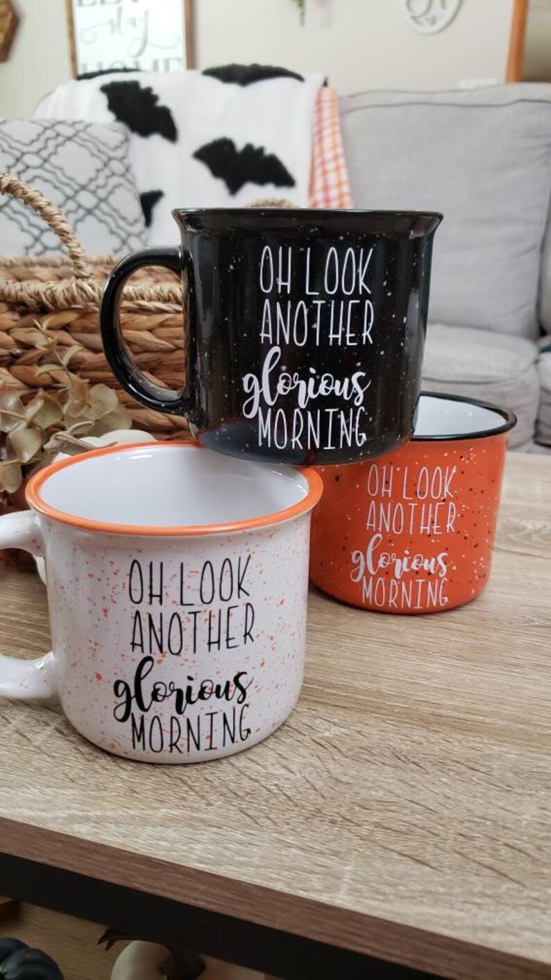 May include: Three ceramic mugs with the text "OH LOOK ANOTHER glorious MORNING". The mugs are black, orange, and white with speckled designs. The white mug has an orange rim, and the black mug has a black handle.