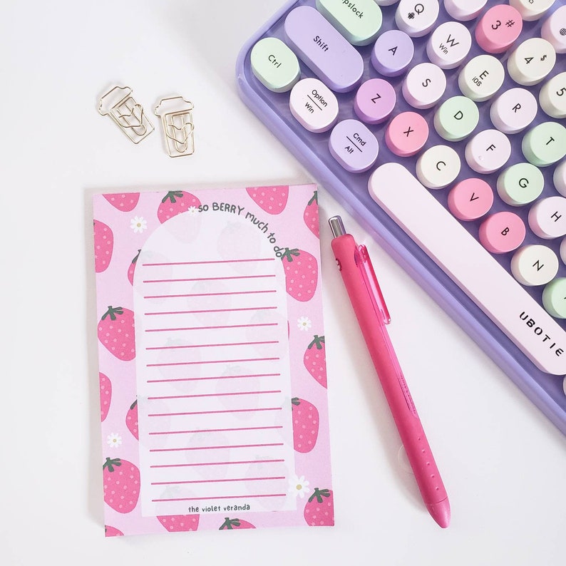 May include: A pink notepad with a strawberry pattern and the text "so BERRY much to do" sits next to a pink pen and a pastel-colored keyboard. Two gold-colored coffee cup paper clips are also visible.