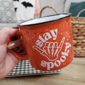 May include: Orange ceramic mug with a white speckled glaze. The mug has a white design of a skeleton hand making a shaka sign with the text "Stay Spooky" on it.