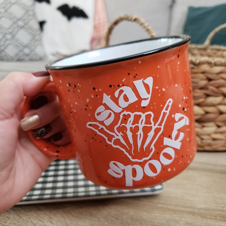 May include: Orange ceramic mug with a white speckled glaze. The mug has a white design of a skeleton hand making a shaka sign with the text "Stay Spooky" on it.