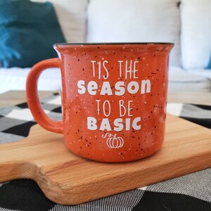 May include: Orange ceramic mug with black speckles and a white rim. The mug has the text "Tis the season to be basic" and a small pumpkin graphic.