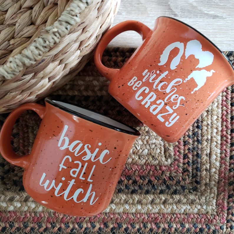 May include: Two orange ceramic mugs with white text. One mug says "basic fall witch" and the other says "witches be crazy".