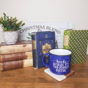 Puede incluir: Una taza de cer&aacute;mica azul con texto blanco que dice "books because it's too peopley outside". La taza est&aacute; sobre un posavasos de madera y una pila de libros.