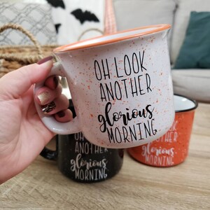 May include: White speckled ceramic mug with an orange rim and the text "OH LOOK ANOTHER glorious MORNING" in black. The mug is held by a person. Two other mugs, one black and one orange, are visible.