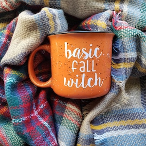 May include: Orange ceramic mug with a black rim and handle. The mug has white speckled paint and the words "basic fall witch" written in white.