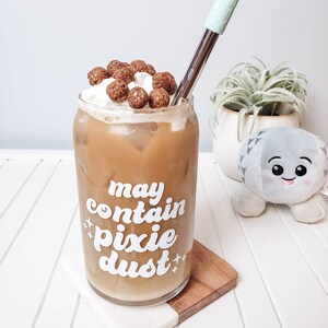 Puede incluir: Un vaso de café helado con crema batida y cereales encima. El vaso tiene el texto "may contain pixie dust" impreso.