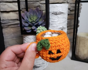 May include: Crocheted orange pumpkin mug with a green leaf and a smiling jack-o'-lantern face. The mug is filled with white marshmallows and brown liquid. A festive Halloween decoration.