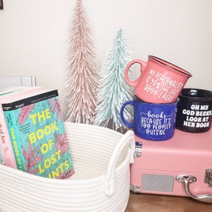 Puede incluir: Tres tazas con frases humor&iacute;sticas sobre libros. Las tazas est&aacute;n apiladas una encima de la otra. La taza de arriba es rosa con letras negras, la taza del medio es azul con letras blancas y la taza de abajo es negra con letras blancas. Las tazas est&aacute;n sobre una maleta rosa con una cesta de cuerda blanca delante.