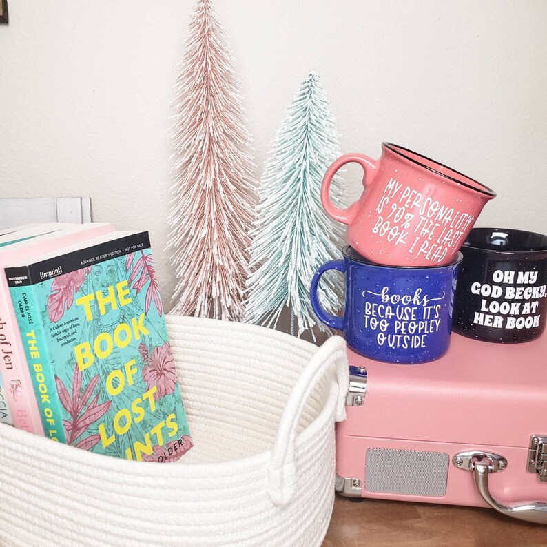 Puede incluir: Tres tazas con frases humor&iacute;sticas sobre libros. Las tazas est&aacute;n apiladas una encima de la otra. La taza de arriba es rosa con letras negras, la taza del medio es azul con letras blancas y la taza de abajo es negra con letras blancas. Las tazas est&aacute;n sobre una maleta rosa con una cesta de cuerda blanca delante.